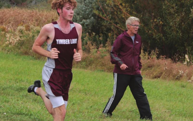 Timber Lake senior Ethan Farlee runs the homestretch of the Panthers’ home meet while coach Donna Aberle encourages him. Photo by Jon Flatland