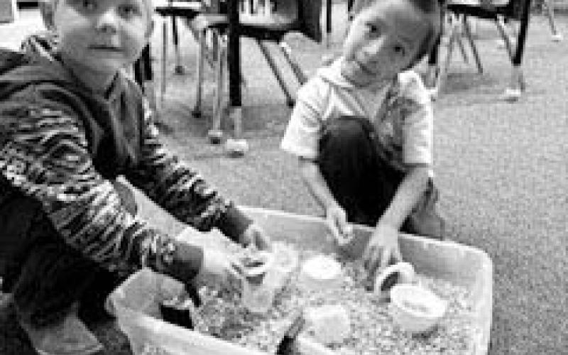 Ryder Schrempp and Kyro Red Bear are working in a corn sensory bin using funnels, cups, bowls, measuring cups and scoops.