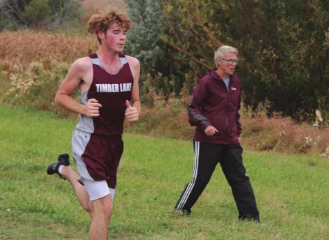 Timber Lake senior Ethan Farlee runs the homestretch of the Panthers’ home meet while coach Donna Aberle encourages him. Photo by Jon Flatland